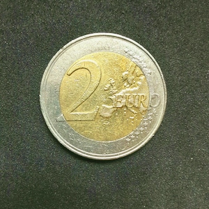 May include: A two-euro coin featuring a bimetallic design with a gold-coloured centre and a silver-coloured outer ring. The centre displays the large numeral 2, the text EURO, and a map of Europe. The outer ring shows stars and lines.