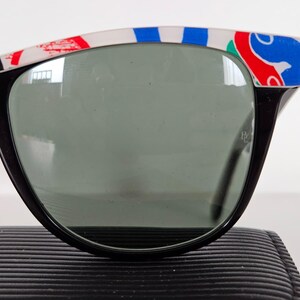 May include: Black sunglasses with a decorative white, red, and blue pattern on the top frame. The lenses are a dark grey colour. The sunglasses are resting on a black, textured surface.