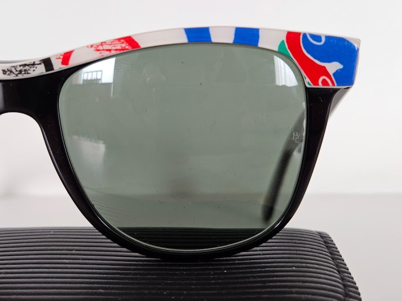 May include: Black sunglasses with a decorative white, red, and blue pattern on the top frame. The lenses are a dark grey colour. The sunglasses are resting on a black, textured surface.