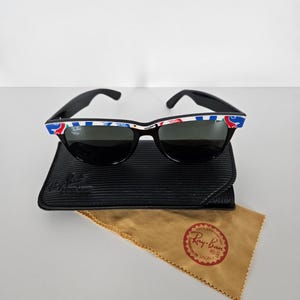 May include: Black sunglasses with a patterned design on the top frame, on a black case and a tan cleaning cloth. The sunglasses have dark lenses and a classic frame shape. The cleaning cloth has a red and gold logo.
