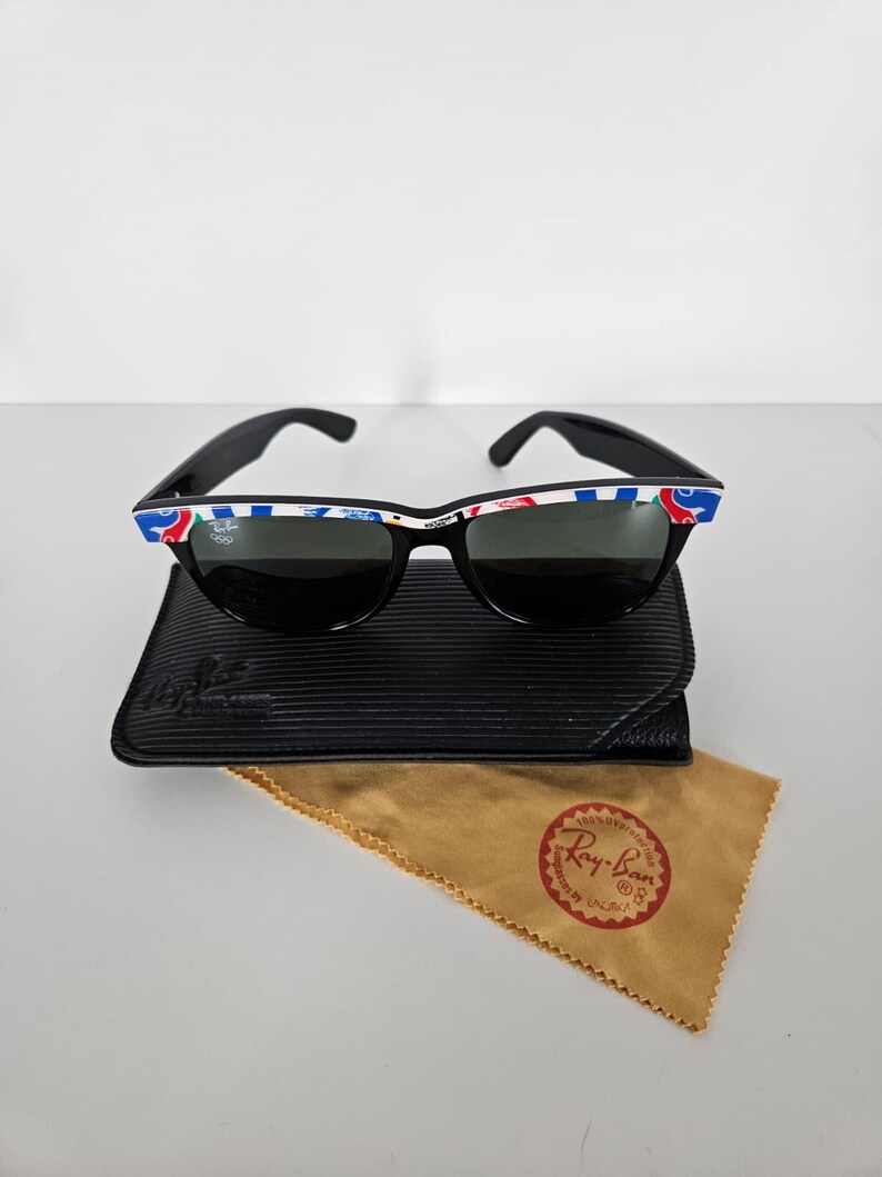 May include: Black sunglasses with a patterned design on the top frame, on a black case and a tan cleaning cloth. The sunglasses have dark lenses and a classic frame shape. The cleaning cloth has a red and gold logo.