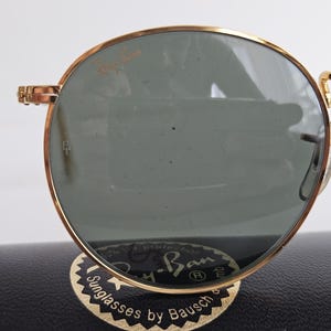 May include: Gold-rimmed Ray-Ban sunglasses with green lenses. The sunglasses are resting on a black case with the Ray-Ban logo and the text "Sunglasses by Bausch". The frames have a classic round shape.