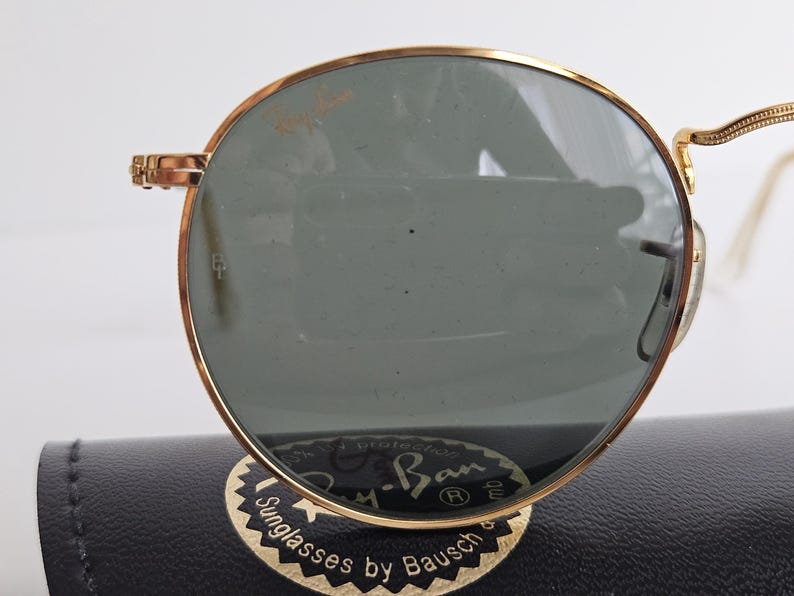 May include: Gold-rimmed Ray-Ban sunglasses with green lenses. The sunglasses are resting on a black case with the Ray-Ban logo and the text "Sunglasses by Bausch". The frames have a classic round shape.