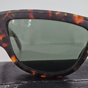 May include: Close-up of sunglasses with a tortoiseshell pattern frame and green lenses. The sunglasses have a thick, angular design. The frame features shades of brown and orange.