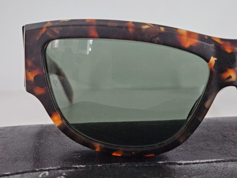 May include: Close-up of sunglasses with a tortoiseshell pattern frame and green lenses. The sunglasses have a thick, angular design. The frame features shades of brown and orange.