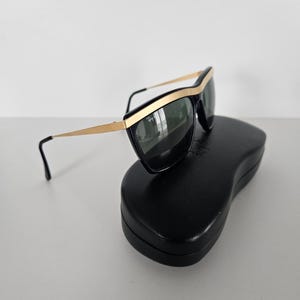 Puede incluir: Gafas de sol negras con detalles dorados y lentes oscuras sobre un estuche negro. Las gafas tienen un dise&ntilde;o elegante y moderno con montura dorada y varillas negras. El estuche es de forma rectangular simple.