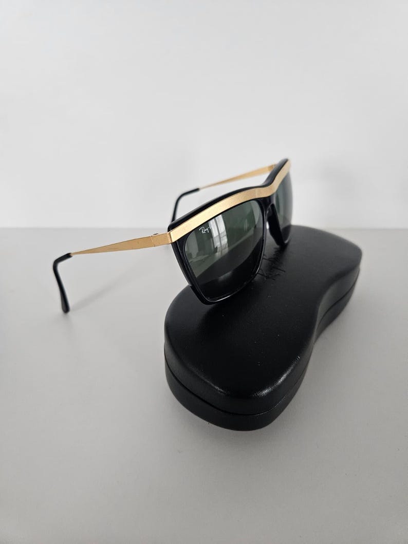 Puede incluir: Gafas de sol negras con detalles dorados y lentes oscuras sobre un estuche negro. Las gafas tienen un dise&ntilde;o elegante y moderno con montura dorada y varillas negras. El estuche es de forma rectangular simple.