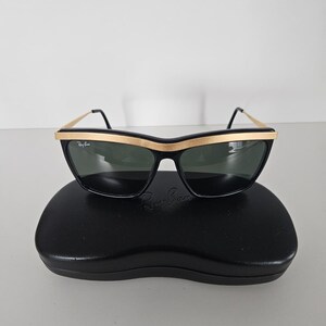 Puede incluir: Gafas de sol negras con montura dorada y lentes oscuras. Las gafas de sol est&aacute;n sobre un estuche negro. El logotipo de Ray-Ban es visible en las lentes y el estuche. Dise&ntilde;o retro y cl&aacute;sico.
