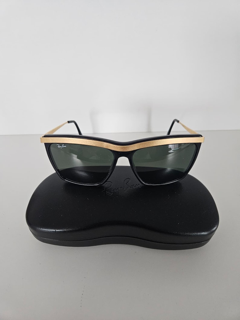 Puede incluir: Gafas de sol negras con montura dorada y lentes oscuras. Las gafas de sol est&aacute;n sobre un estuche negro. El logotipo de Ray-Ban es visible en las lentes y el estuche. Dise&ntilde;o retro y cl&aacute;sico.