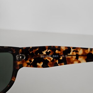 May include: Close-up of sunglasses with a tortoiseshell pattern frame and dark lenses. The text "B&L RAY-BAN S.A." is printed on the arm of the sunglasses. The sunglasses have a classic, rectangular shape.