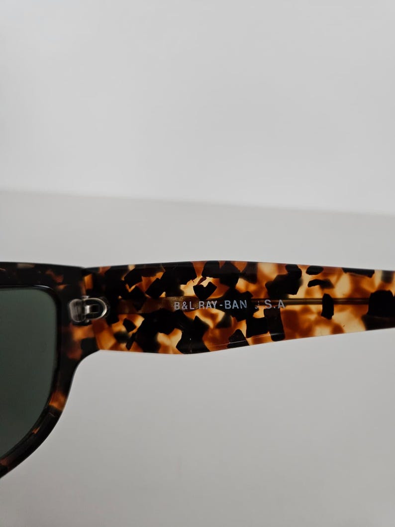 May include: Close-up of sunglasses with a tortoiseshell pattern frame and dark lenses. The text "B&L RAY-BAN S.A." is printed on the arm of the sunglasses. The sunglasses have a classic, rectangular shape.