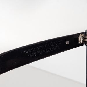 May include: Close-up of a black sunglasses arm. The text "SPORT WAYFARER II 1992 BARCELONA" is printed in white on the arm. The sunglasses arm is angled, showing a hinge and part of the frame against a white background.