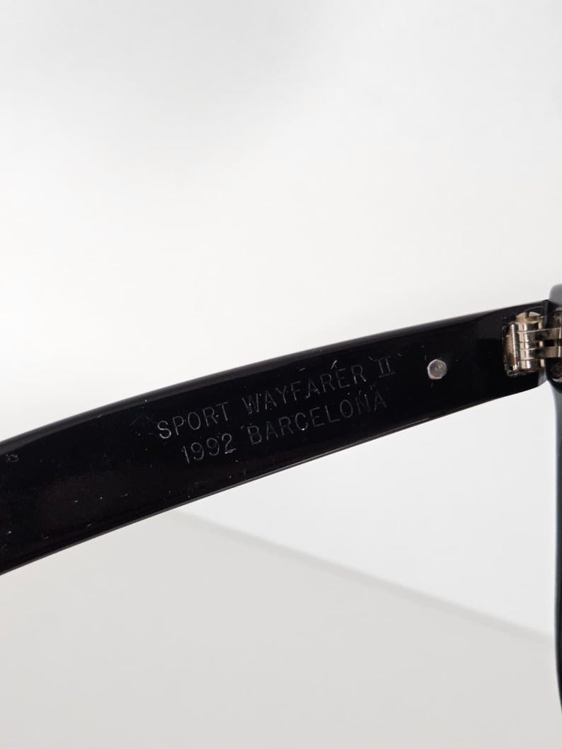 May include: Close-up of a black sunglasses arm. The text "SPORT WAYFARER II 1992 BARCELONA" is printed in white on the arm. The sunglasses arm is angled, showing a hinge and part of the frame against a white background.