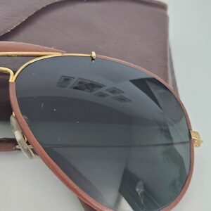 Puede incluir: Gafas de sol estilo aviador con lentes oscuras y montura marr&oacute;n. Las gafas tienen detalles dorados y se combinan con un estuche de cuero marr&oacute;n. Las lentes reflejan el entorno.