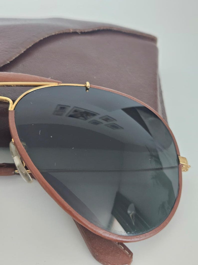 Puede incluir: Gafas de sol estilo aviador con lentes oscuras y montura marr&oacute;n. Las gafas tienen detalles dorados y se combinan con un estuche de cuero marr&oacute;n. Las lentes reflejan el entorno.