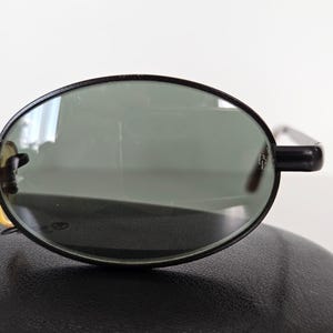 Puede incluir: Gafas de sol ovaladas con montura negra y lentes verdes. Las gafas de sol est&aacute;n colocadas sobre un estuche de cuero negro. Las monturas tienen un dise&ntilde;o cl&aacute;sico y las lentes parecen estar en buen estado.