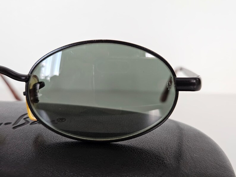 Puede incluir: Gafas de sol ovaladas con montura negra y lentes verdes. Las gafas de sol est&aacute;n colocadas sobre un estuche de cuero negro. Las monturas tienen un dise&ntilde;o cl&aacute;sico y las lentes parecen estar en buen estado.