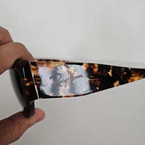 May include: Tortoiseshell-patterned sunglasses with the Ray-Ban logo visible on the temple. The sunglasses have a dark brown frame and lenses. The image is taken from a side angle.