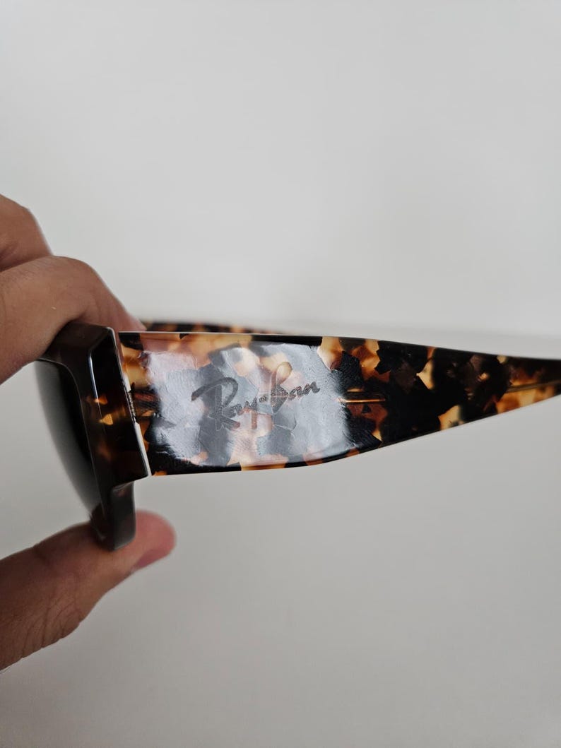 May include: Tortoiseshell-patterned sunglasses with the Ray-Ban logo visible on the temple. The sunglasses have a dark brown frame and lenses. The image is taken from a side angle.