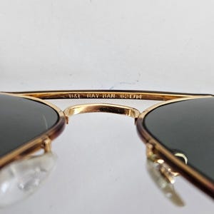 Puede incluir: Primer plano de gafas de sol Ray-Ban con montura dorada y lentes oscuras. El texto "B&L RAY-BAN 62[]14" es visible en la montura. Las gafas de sol tienen un dise&ntilde;o cl&aacute;sico de aviador.