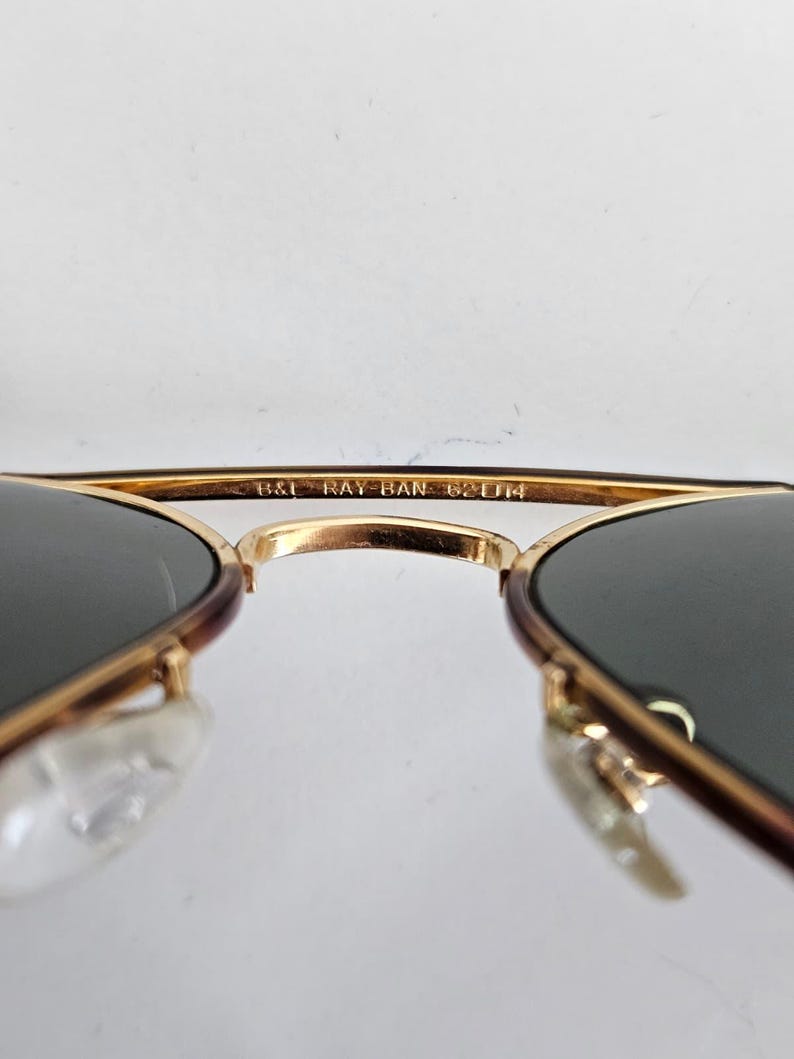Puede incluir: Primer plano de gafas de sol Ray-Ban con montura dorada y lentes oscuras. El texto "B&L RAY-BAN 62[]14" es visible en la montura. Las gafas de sol tienen un dise&ntilde;o cl&aacute;sico de aviador.