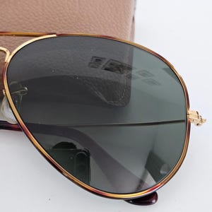 Puede incluir: Gafas de sol estilo aviador con montura de metal dorado y lentes verde oscuro. Las gafas de sol tienen una forma cl&aacute;sica de l&aacute;grima y se combinan con un estuche de cuero marr&oacute;n claro. La montura presenta un acento rojo-marr&oacute;n.