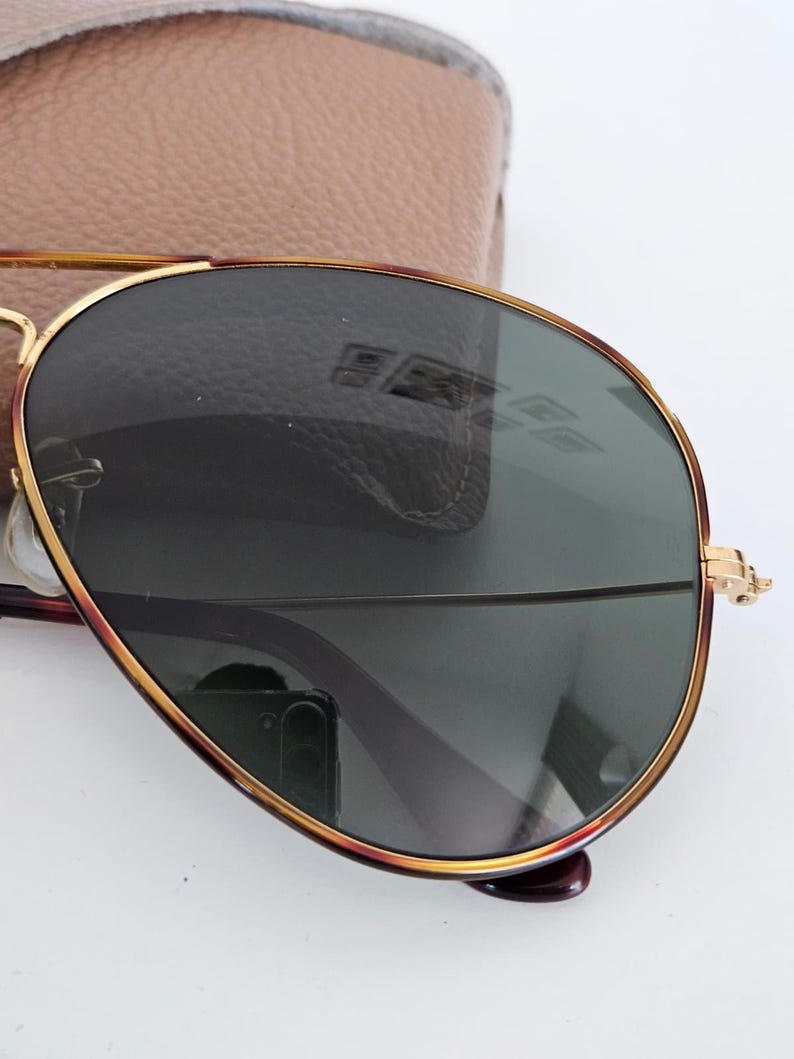 Puede incluir: Gafas de sol estilo aviador con montura de metal dorado y lentes verde oscuro. Las gafas de sol tienen una forma cl&aacute;sica de l&aacute;grima y se combinan con un estuche de cuero marr&oacute;n claro. La montura presenta un acento rojo-marr&oacute;n.