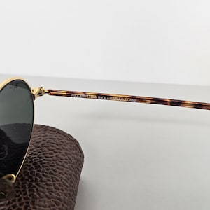 May include: Gold-rimmed sunglasses with dark lenses and tortoiseshell arms. The text "RAY-BAN USA BY BAUSCH & LOMB" is printed on the arm. The sunglasses are resting on a brown leather case.