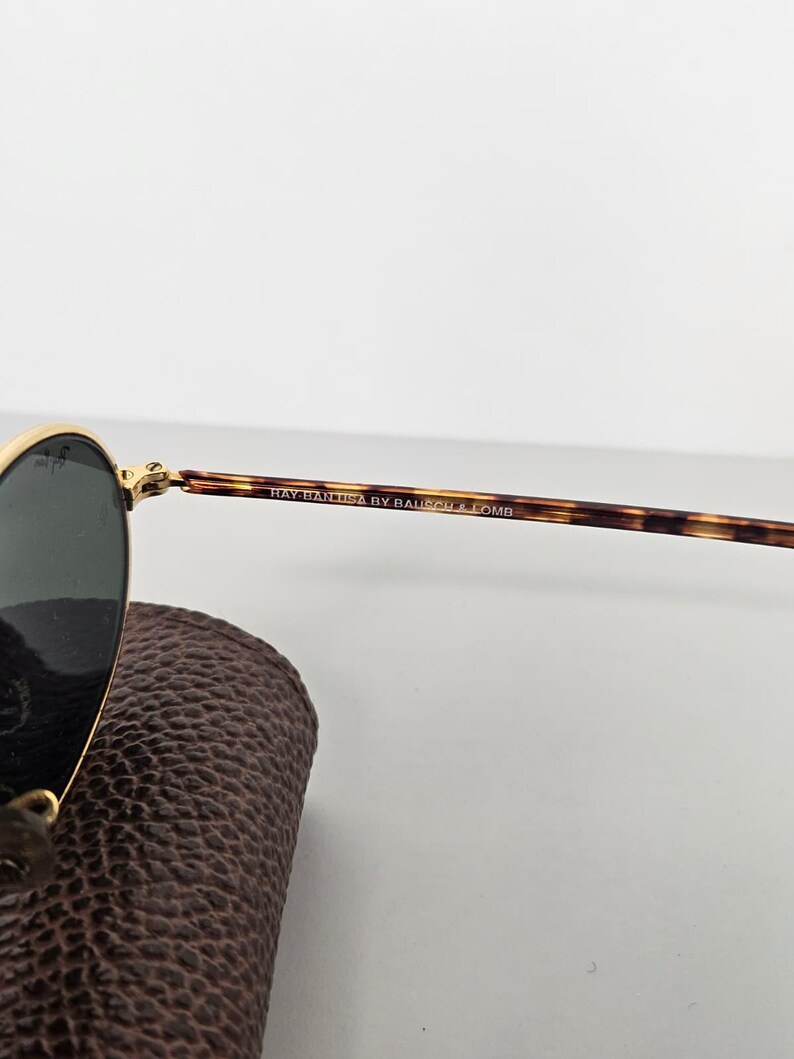 May include: Gold-rimmed sunglasses with dark lenses and tortoiseshell arms. The text "RAY-BAN USA BY BAUSCH & LOMB" is printed on the arm. The sunglasses are resting on a brown leather case.