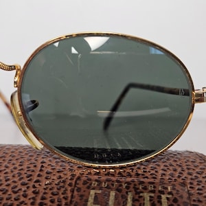 May include: Oval sunglasses with gold frames and dark green lenses. The frames have a detailed design, and the temples are slender. The sunglasses are on a brown leather case.