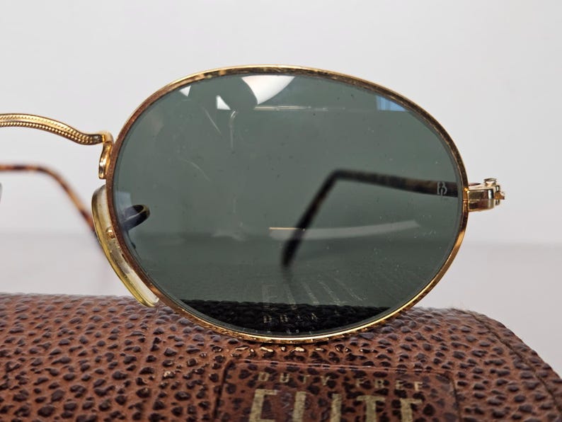 May include: Oval sunglasses with gold frames and dark green lenses. The frames have a detailed design, and the temples are slender. The sunglasses are on a brown leather case.