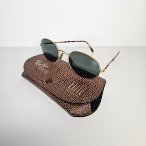 May include: Oval sunglasses with gold frames and dark lenses, presented on a brown leather case. The case features a textured finish and gold lettering that reads "Ray-Ban Sunglasses" and "ELITE". The temples of the sunglasses have a tortoiseshell pattern.