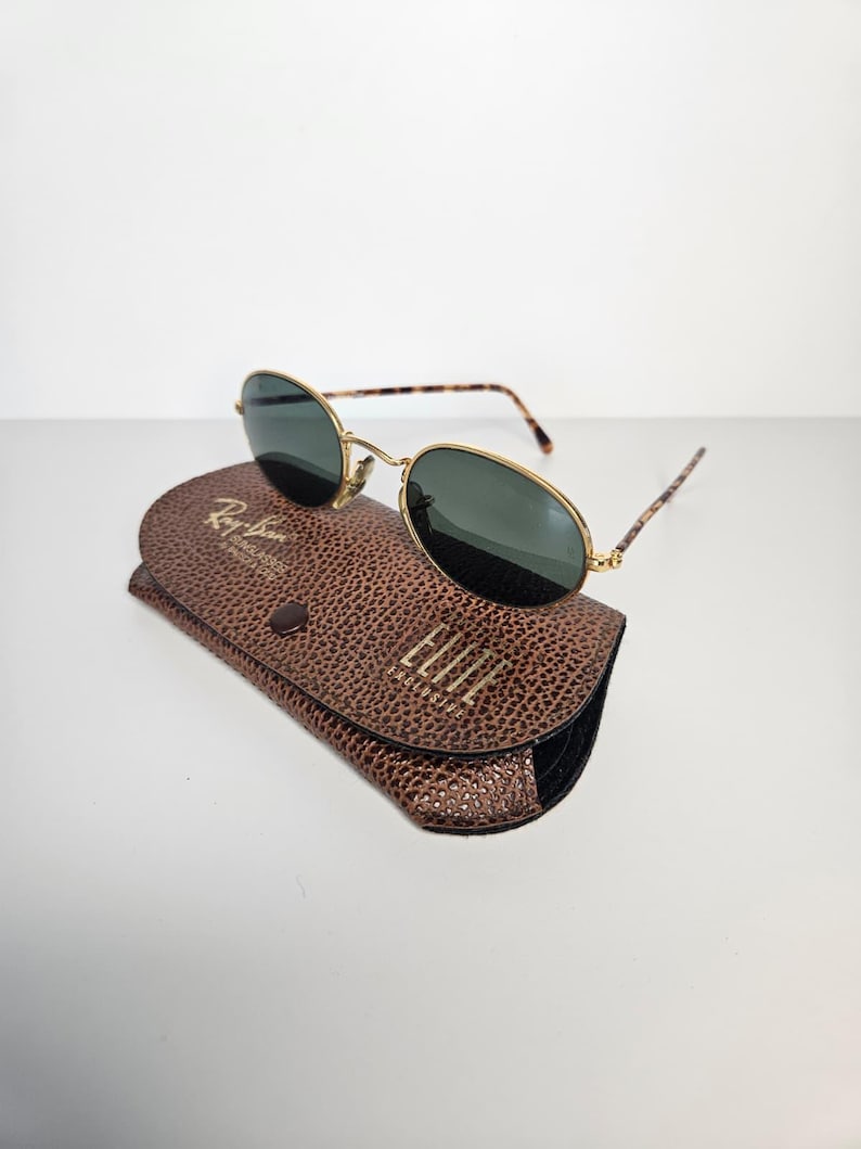May include: Oval sunglasses with gold frames and dark lenses, presented on a brown leather case. The case features a textured finish and gold lettering that reads "Ray-Ban Sunglasses" and "ELITE". The temples of the sunglasses have a tortoiseshell pattern.