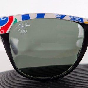 May include: Black Ray-Ban sunglasses with a colourful pattern on the top frame. The lenses are a dark grey colour. The Ray-Ban logo and Olympic rings are visible on the lens.