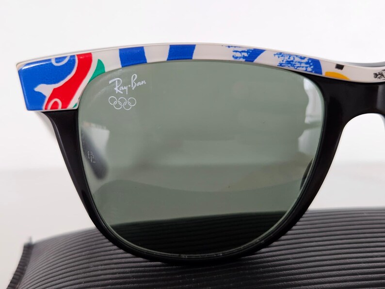 May include: Black Ray-Ban sunglasses with a colourful pattern on the top frame. The lenses are a dark grey colour. The Ray-Ban logo and Olympic rings are visible on the lens.