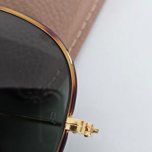 Puede incluir: Primer plano de gafas de sol con montura dorada y lentes oscuras. La montura tiene un patr&oacute;n de carey en la parte superior y los laterales. La bisagra es dorada y las lentes son de color verde oscuro. Un estuche marr&oacute;n claro est&aacute; en el fondo.