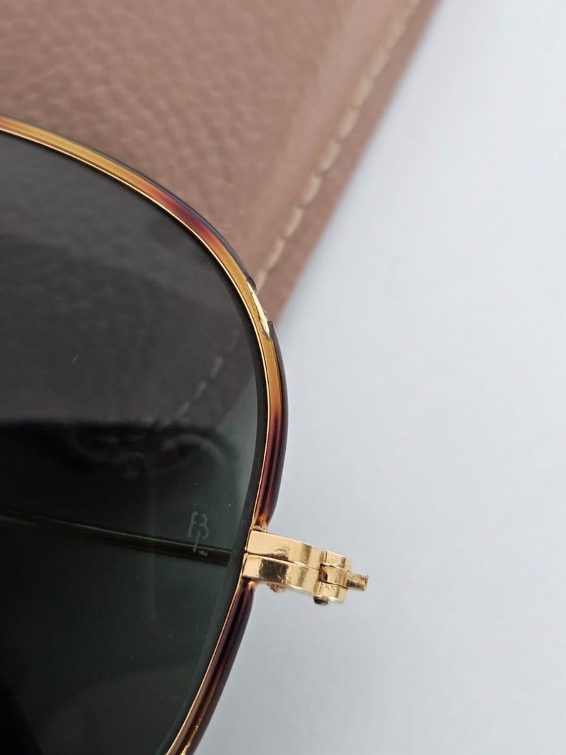Puede incluir: Primer plano de gafas de sol con montura dorada y lentes oscuras. La montura tiene un patr&oacute;n de carey en la parte superior y los laterales. La bisagra es dorada y las lentes son de color verde oscuro. Un estuche marr&oacute;n claro est&aacute; en el fondo.