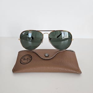 Puede incluir: Gafas de sol aviador con montura dorada y lentes verdes sobre un estuche de cuero marr&oacute;n. Las gafas tienen un dise&ntilde;o cl&aacute;sico con doble puente y varillas curvas. El estuche es rectangular con cierre de bot&oacute;n a presi&oacute;n.