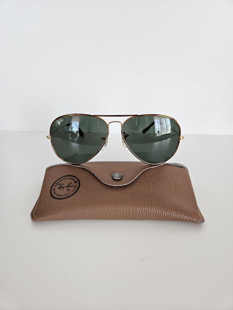 Puede incluir: Gafas de sol aviador con montura dorada y lentes verdes sobre un estuche de cuero marr&oacute;n. Las gafas tienen un dise&ntilde;o cl&aacute;sico con doble puente y varillas curvas. El estuche es rectangular con cierre de bot&oacute;n a presi&oacute;n.