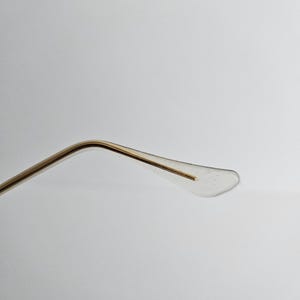 May include: Close-up of the temple arm of eyeglasses. The arm is gold-colored metal, curving towards a clear, plastic earpiece. The earpiece has a small, subtle design. The background is a plain, light gray.