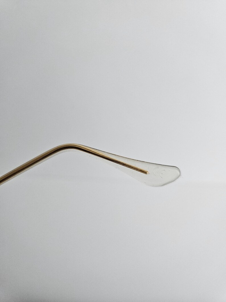 May include: Close-up of the temple arm of eyeglasses. The arm is gold-colored metal, curving towards a clear, plastic earpiece. The earpiece has a small, subtle design. The background is a plain, light gray.