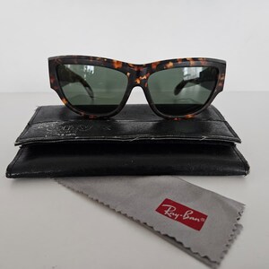 May include: Tortoiseshell sunglasses with green lenses rest on a black leather case. A gray cleaning cloth with a red Ray-Ban logo is below the case. The sunglasses have a thick frame and a classic design.