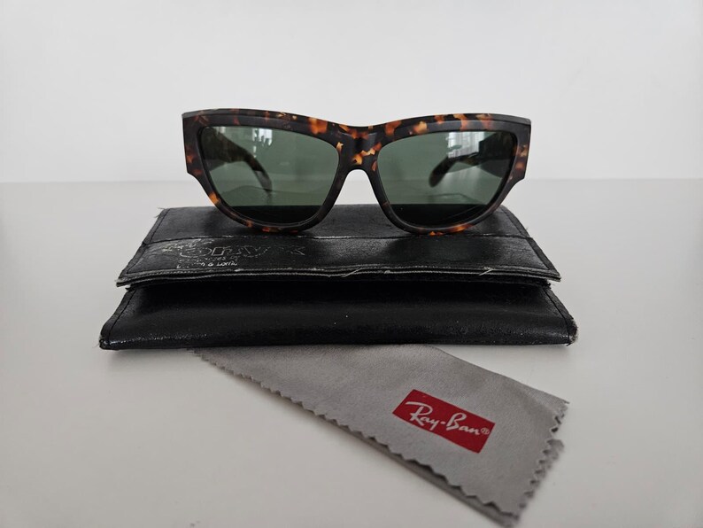 May include: Tortoiseshell sunglasses with green lenses rest on a black leather case. A gray cleaning cloth with a red Ray-Ban logo is below the case. The sunglasses have a thick frame and a classic design.
