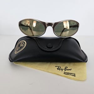 May include: A pair of Ray-Ban sunglasses with a dark metallic frame and green-tinted lenses rests atop a black leather case. A yellow cleaning cloth with the Ray-Ban logo is visible below the case.