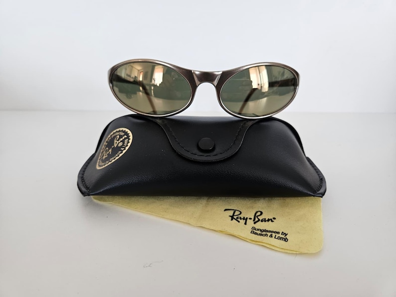 May include: A pair of Ray-Ban sunglasses with a dark metallic frame and green-tinted lenses rests atop a black leather case. A yellow cleaning cloth with the Ray-Ban logo is visible below the case.