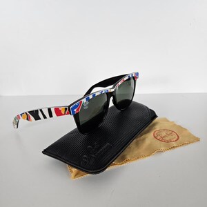 May include: Black sunglasses with a patterned frame, on a black case and a tan cleaning cloth. The sunglasses have dark lenses and a square shape. The case is textured. The cloth has a logo.