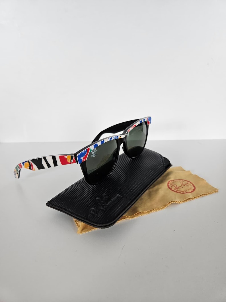 May include: Black sunglasses with a patterned frame, on a black case and a tan cleaning cloth. The sunglasses have dark lenses and a square shape. The case is textured. The cloth has a logo.