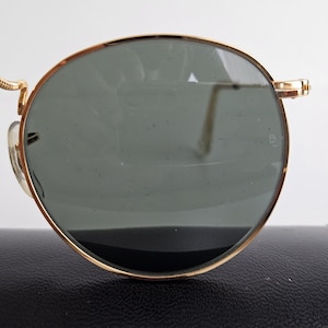 May include: Close-up of gold-rimmed sunglasses with dark green lenses. The round frames have a classic design. The sunglasses are resting on a black case, suggesting a vintage or luxury item.