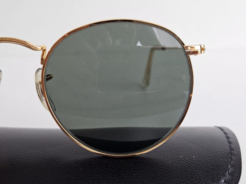 May include: Close-up of gold-rimmed sunglasses with dark green lenses. The round frames have a classic design. The sunglasses are resting on a black case, suggesting a vintage or luxury item.