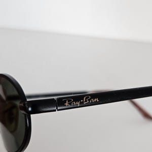 Puede incluir: Primer plano de gafas de sol Ray-Ban negras. Las gafas de sol tienen una montura redonda y lentes oscuras. El logotipo de Ray-Ban es visible en la patilla. La montura es negra con un acento marr&oacute;n en la patilla.